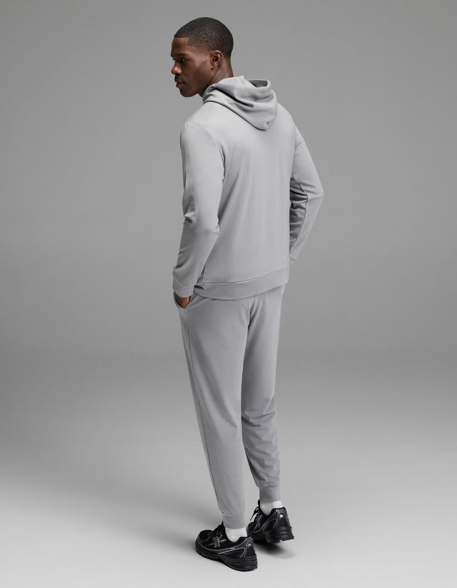 A person models the Cozy Earth Men’s Brushed Bamboo Hoodie & Jogger Set in light gray, hands in pockets, paired with black sneakers and photographed against a plain gray background.