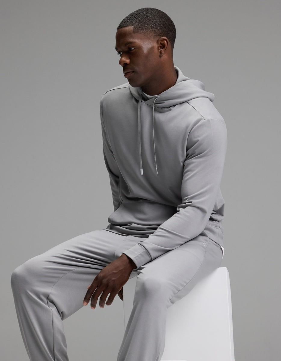 Dressed in the Cozy Earth Men’s Brushed Bamboo Hoodie & Jogger Set, a man sits thoughtfully on a white stool against a plain gray background, his hands resting on his knees as he looks down.
