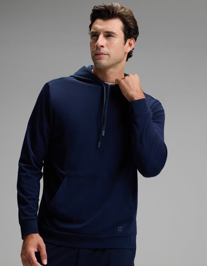 A person wearing the Cozy Earth Men’s Brushed Bamboo Hoodie in dark blue stands against a plain gray background, looking slightly to the side while touching the neckline of the hoodie with one hand.