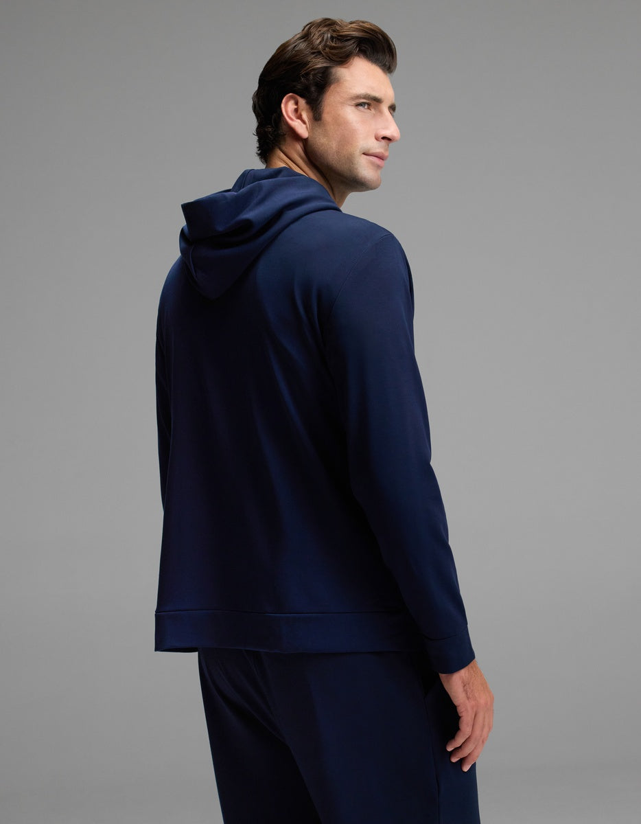 A man with brown hair wears the Cozy Earth Men’s Brushed Bamboo Hoodie in navy blue, paired with matching pants, as he stands facing away and looks over his shoulder against a plain gray background.