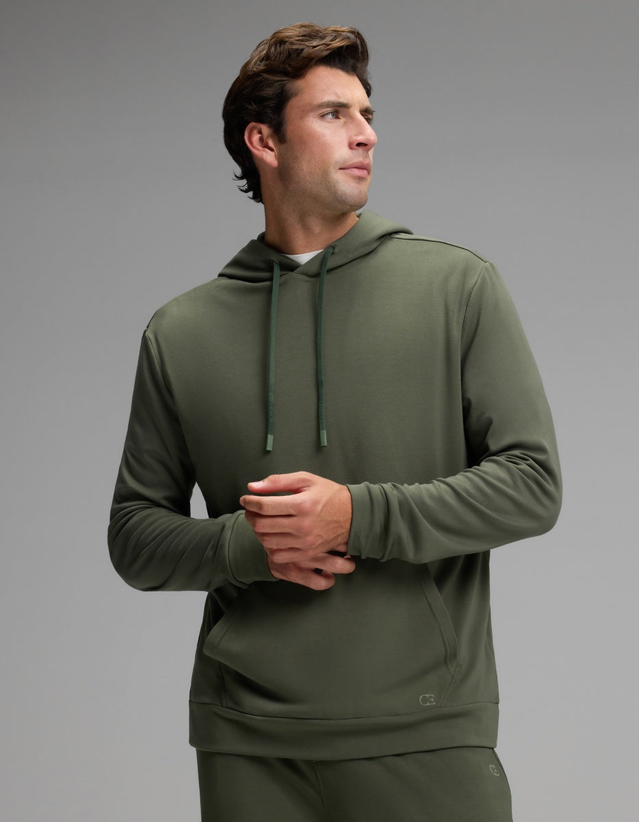 A man stands wearing the Cozy Earth Men’s Brushed Bamboo Hoodie in olive green, paired with matching pants. He looks to his left with his hands clasped, set against a plain light gray background.|Color:Olive