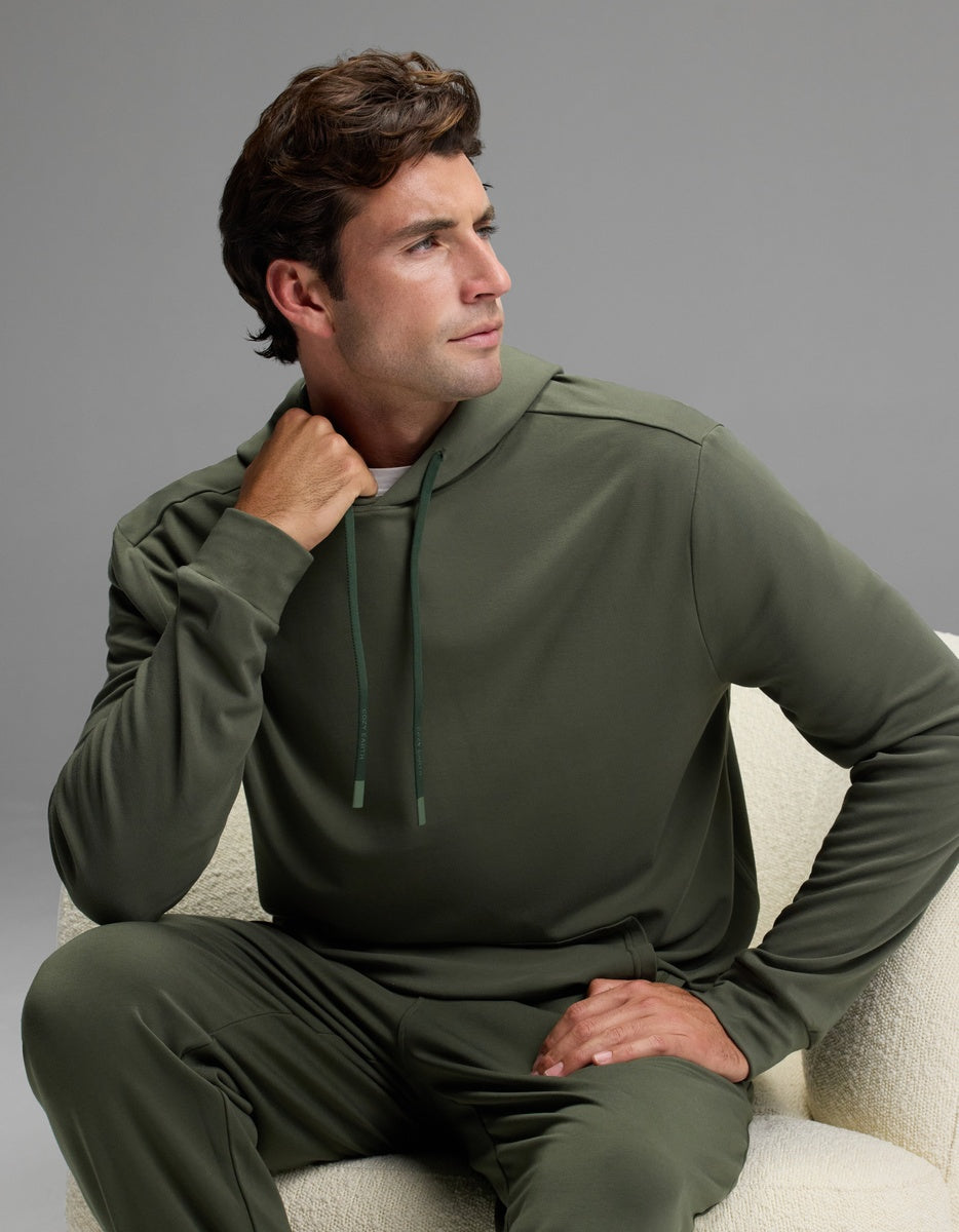 A man in the Cozy Earth Men’s Brushed Bamboo Hoodie and matching pants sits on a light-colored chair, gazing thoughtfully to his left with one hand touching his neck and the other resting on his knee.