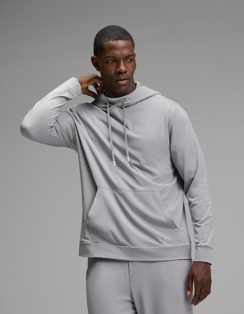 A man wearing the Cozy Earth Men’s Brushed Bamboo Hoodie and matching pants stands against a plain gray background, with one hand touching the back of his neck and the other hanging by his side.|Color:Stone