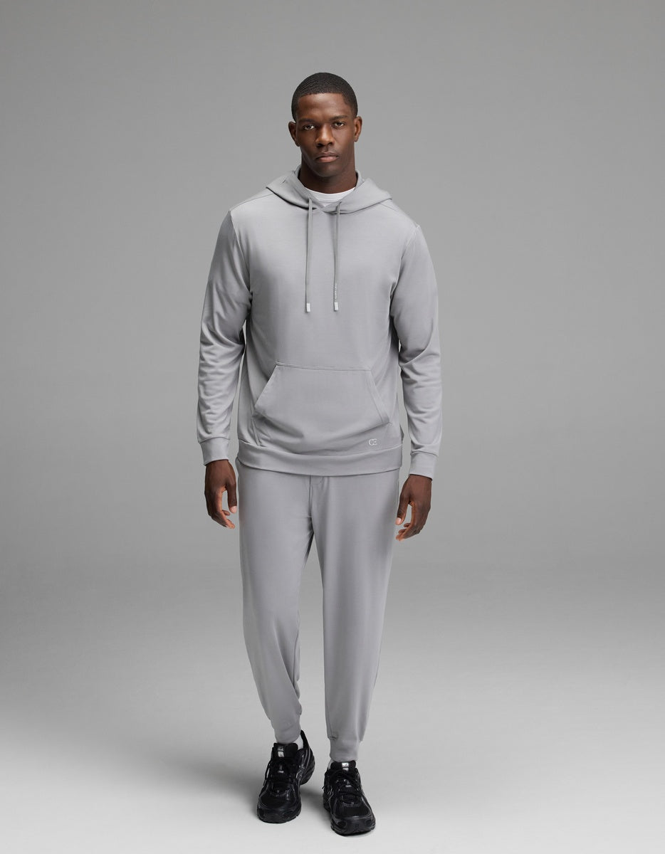 A man wearing the Cozy Earth Men’s Brushed Bamboo Hoodie in light gray and matching pants stands against a plain gray background, looking forward with a neutral expression. He is also wearing black sneakers.