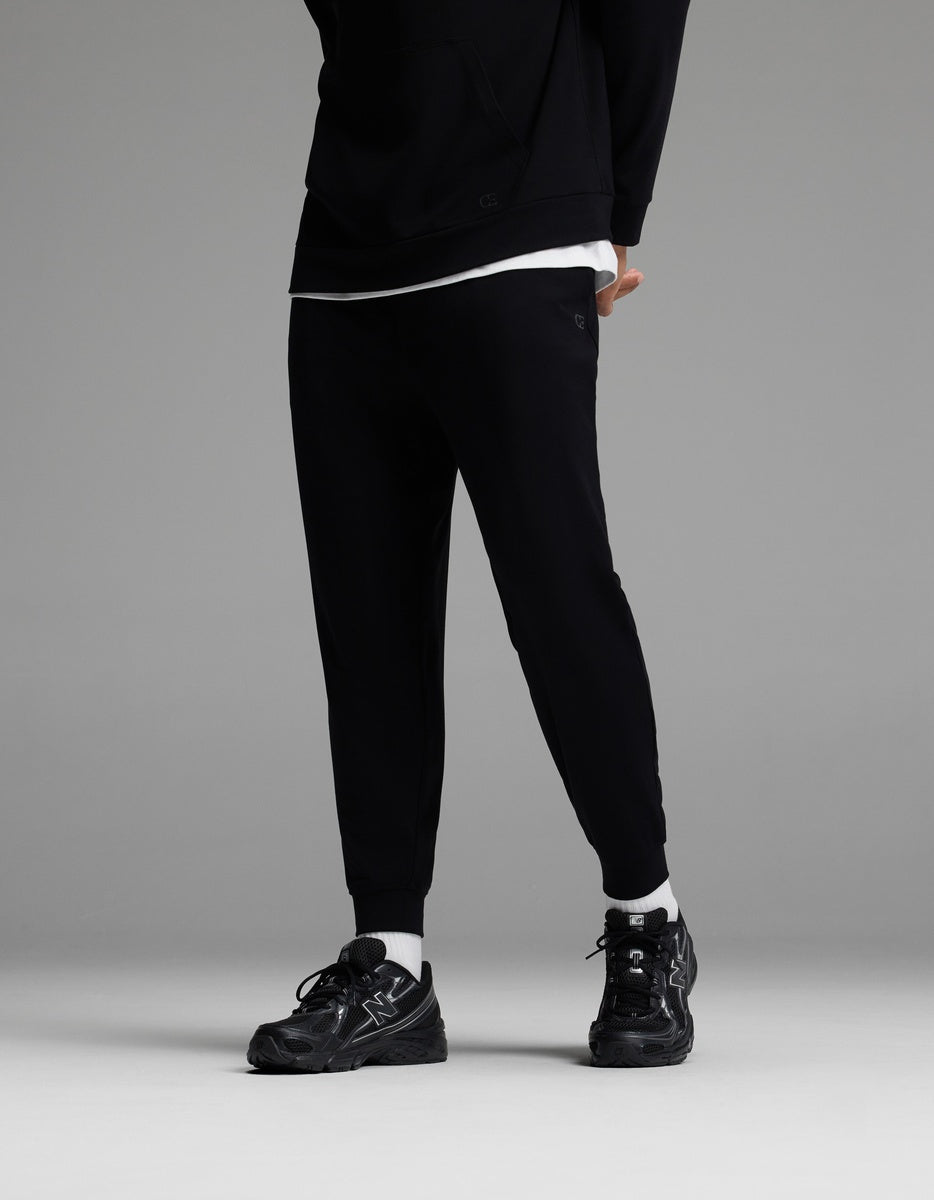 A person is shown from the shoulders down, standing against a gray background and wearing Cozy Earth’s Men’s Brushed Bamboo Joggers in black, paired with a black hoodie, white T-shirt, white socks, and black sneakers. The head is not visible in the image.