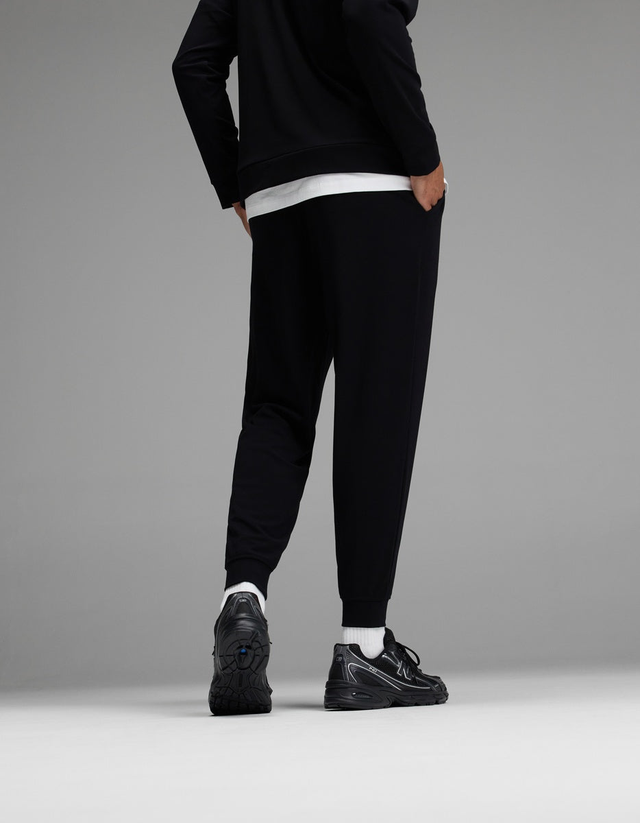 A person stands facing away, wearing black jogger pants, a black jacket, a white shirt, white socks, and black sneakers against a gray background.