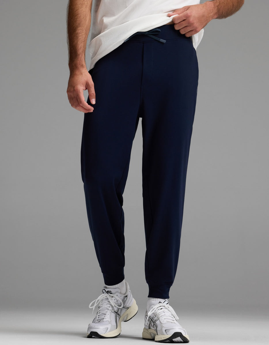 A person stands against a plain gray background, wearing Cozy Earth Men’s Brushed Bamboo Joggers in navy blue, along with a white T-shirt and white athletic sneakers. Only the lower half of the body is visible.|Color:Navy