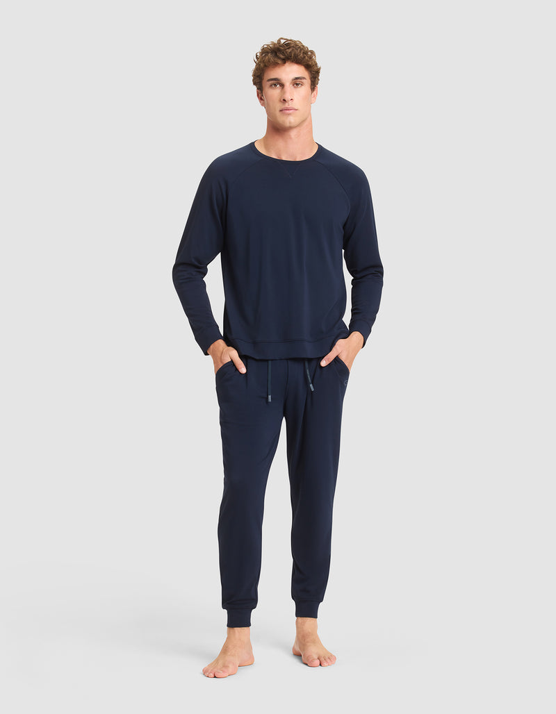 A young man stands barefoot against a light gray background, wearing Cozy Earth's Men's Brushed Bamboo Jogger Pant in navy blue with a matching long-sleeve sweatshirt and his hands in his pockets.