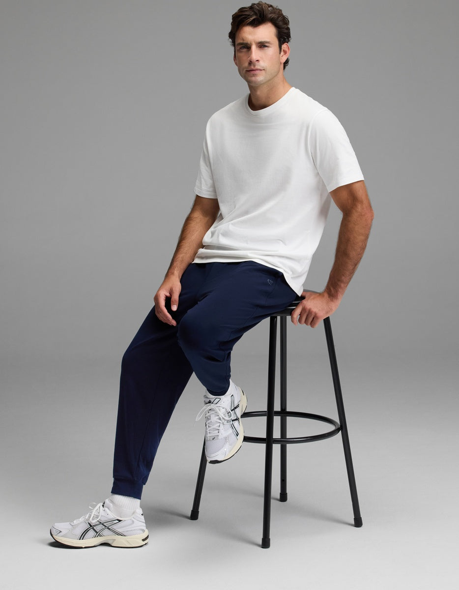 A man sits on a tall black stool wearing a plain white t-shirt, Cozy Earth Men’s Brushed Bamboo Joggers in navy, and white sneakers against a plain gray background.