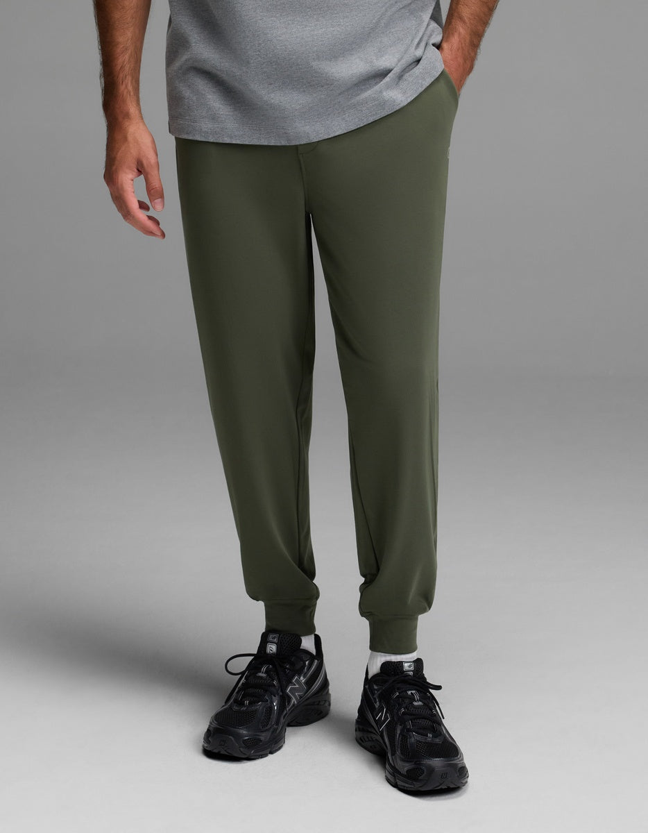 A person wearing Cozy Earth Men’s Brushed Bamboo Joggers in olive green, paired with black sneakers and a gray T-shirt, stands against a plain gray background. Only the lower half of their body is visible.