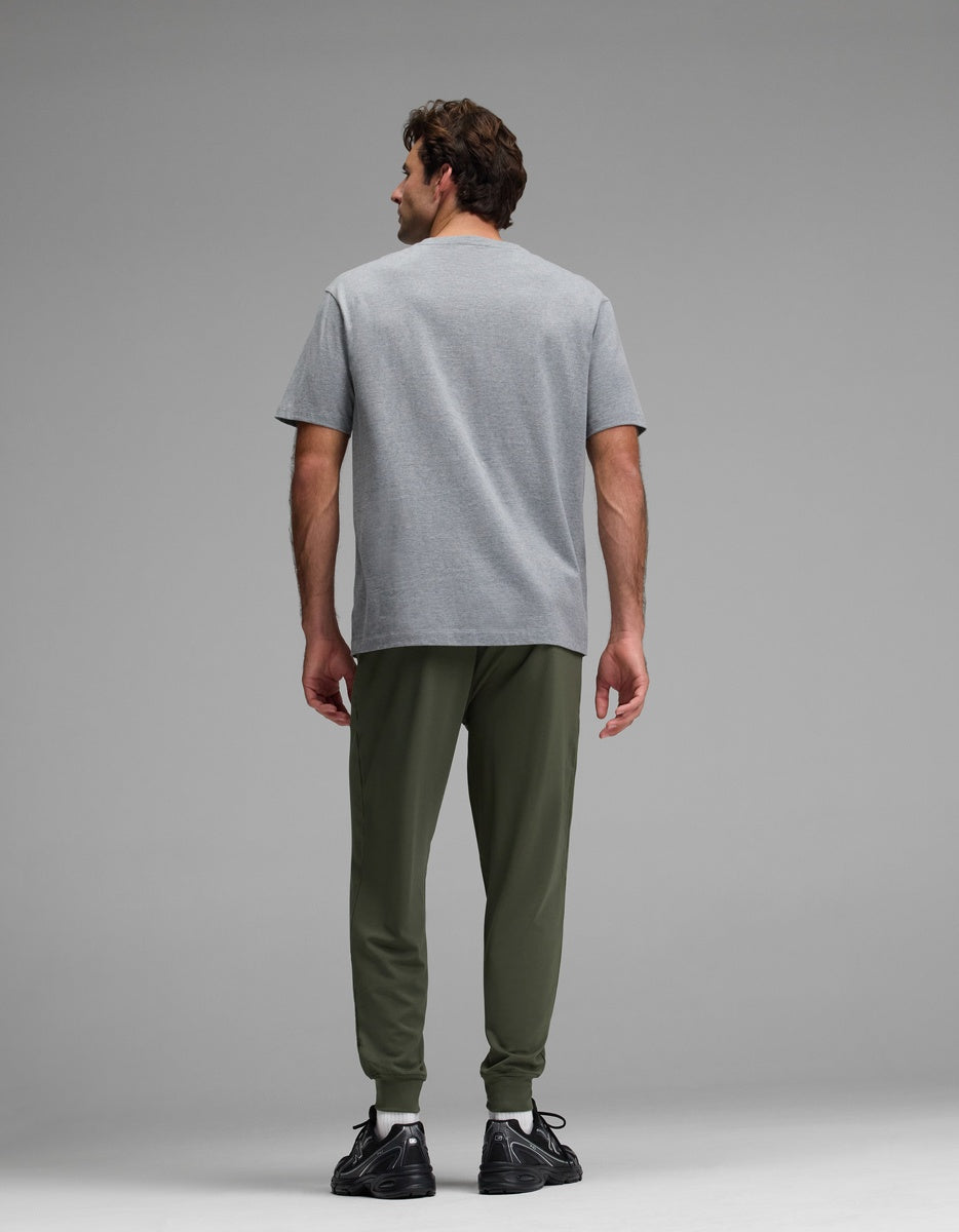 A man stands facing away, wearing Cozy Earth’s Men’s Brushed Bamboo Joggers in olive green, paired with a gray t-shirt and black sneakers against a plain gray background.