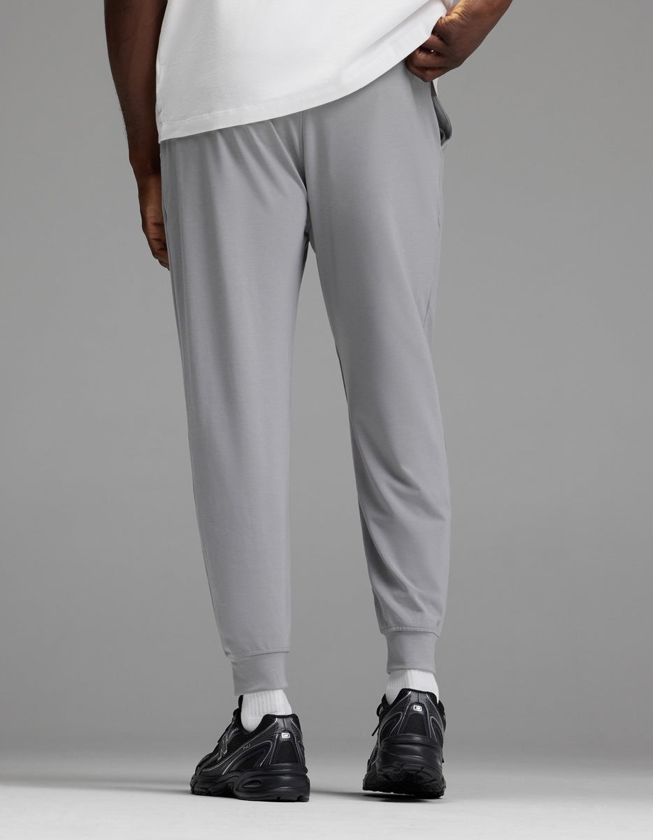 A person is shown from the back, standing against a plain gray background, wearing Cozy Earth Men’s Brushed Bamboo Joggers in light gray paired with a white shirt and black sneakers.
