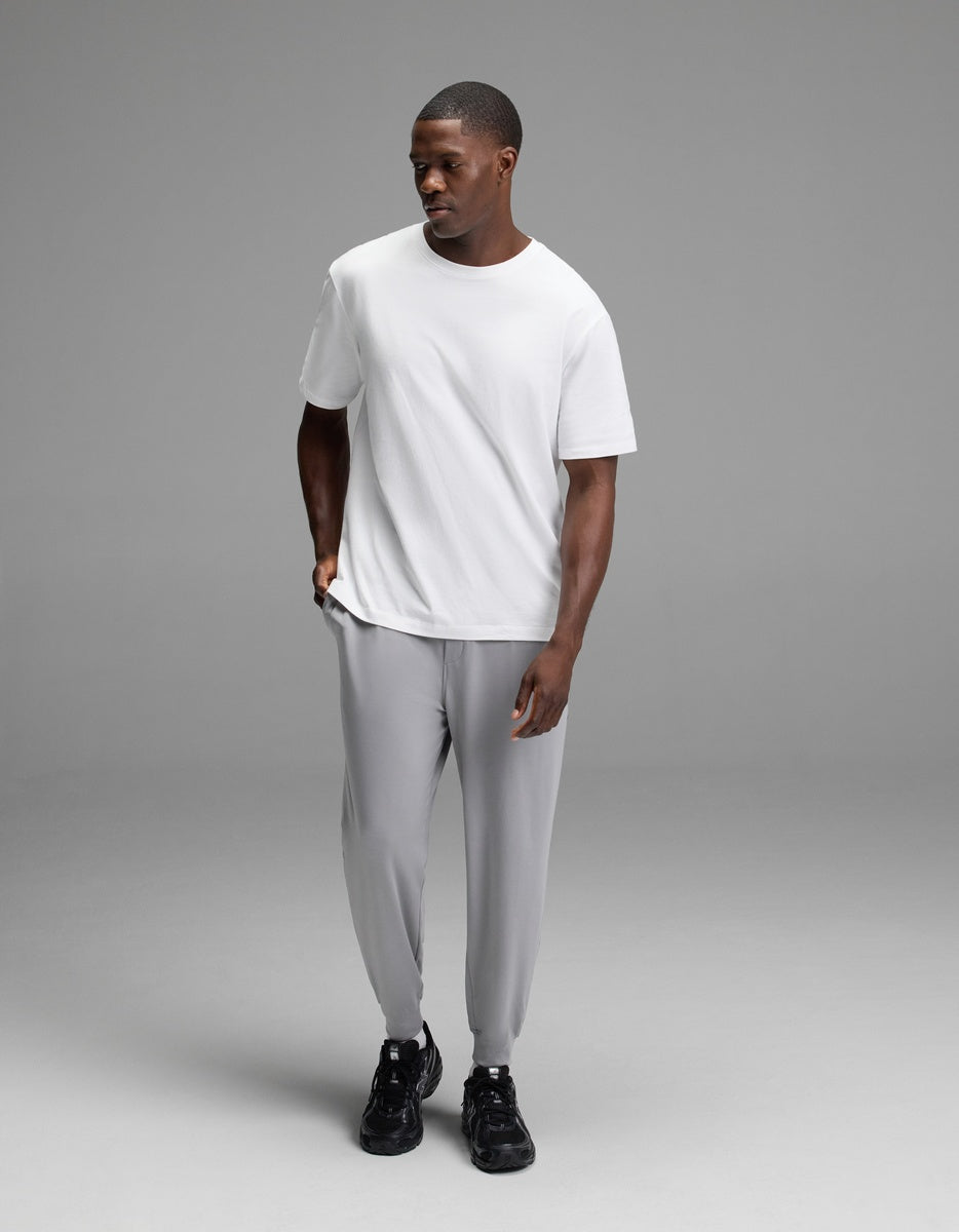 A man wearing Cozy Earth Men’s Brushed Bamboo Joggers in light gray, paired with a plain white T-shirt and black sneakers, stands against a simple gray background, looking slightly down and to the side with one hand in his pocket.