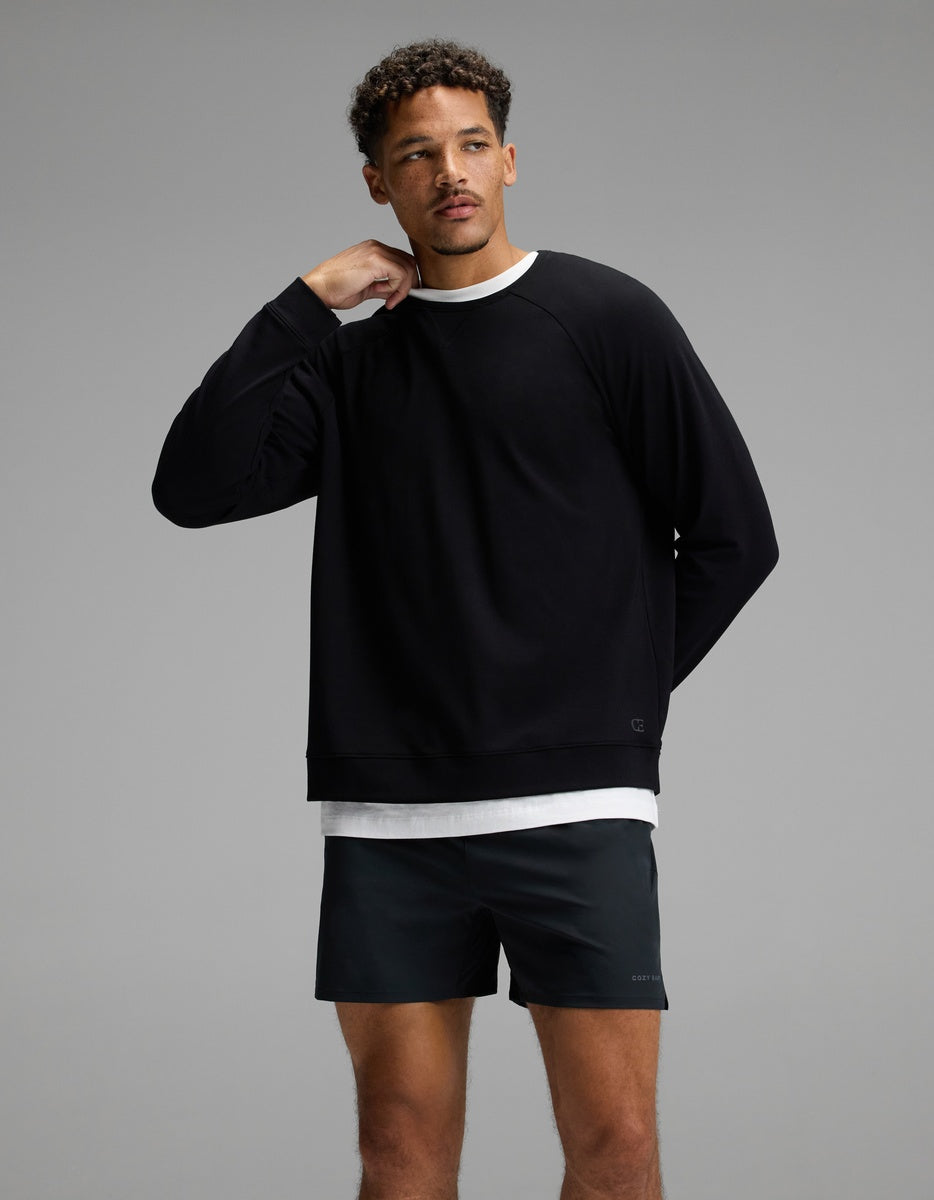 A man stands against a gray background, wearing the Cozy Earth Men’s Brushed Bamboo Pullover Crew in black over a white shirt and black shorts. He has short curly hair and is touching his neck with his right hand, looking slightly to the side.