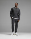 A man stands against a plain gray background, wearing Cozy Earth’s Men’s Brushed Bamboo Jogger Set in dark gray over a white layered shirt and white sneakers, with his hands in his pockets.|Color:Charcoal