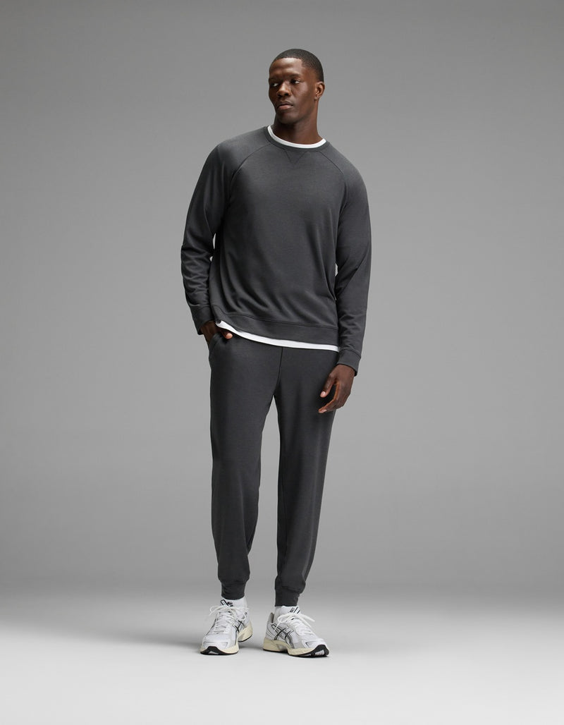A man stands against a plain gray background, wearing Cozy Earth’s Men’s Brushed Bamboo Jogger Set in dark gray over a white layered shirt and white sneakers, with his hands in his pockets.