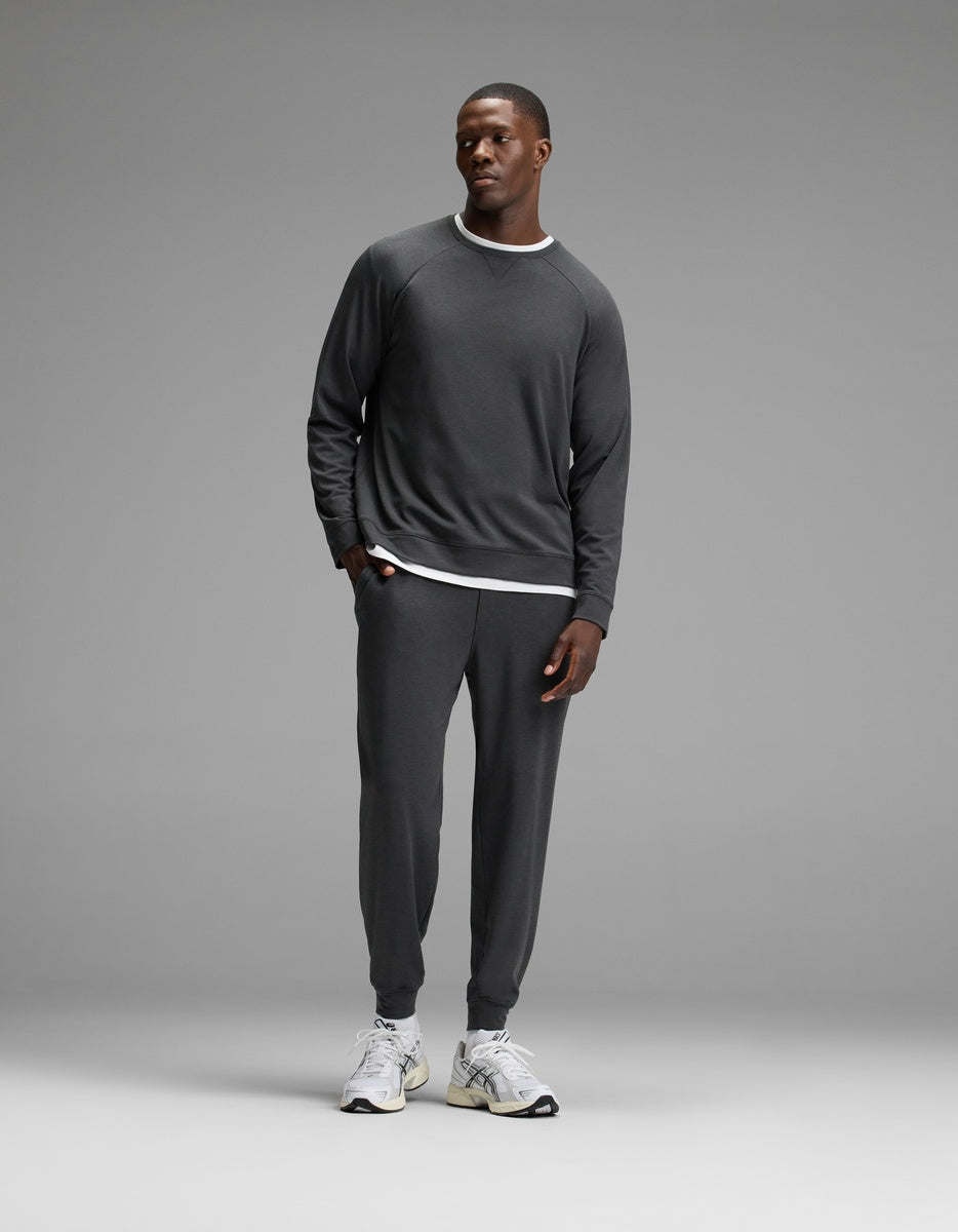 A man stands against a plain gray background, wearing Cozy Earth’s Men’s Brushed Bamboo Jogger Set in dark gray over a white layered shirt and white sneakers, with his hands in his pockets.|Color:Charcoal