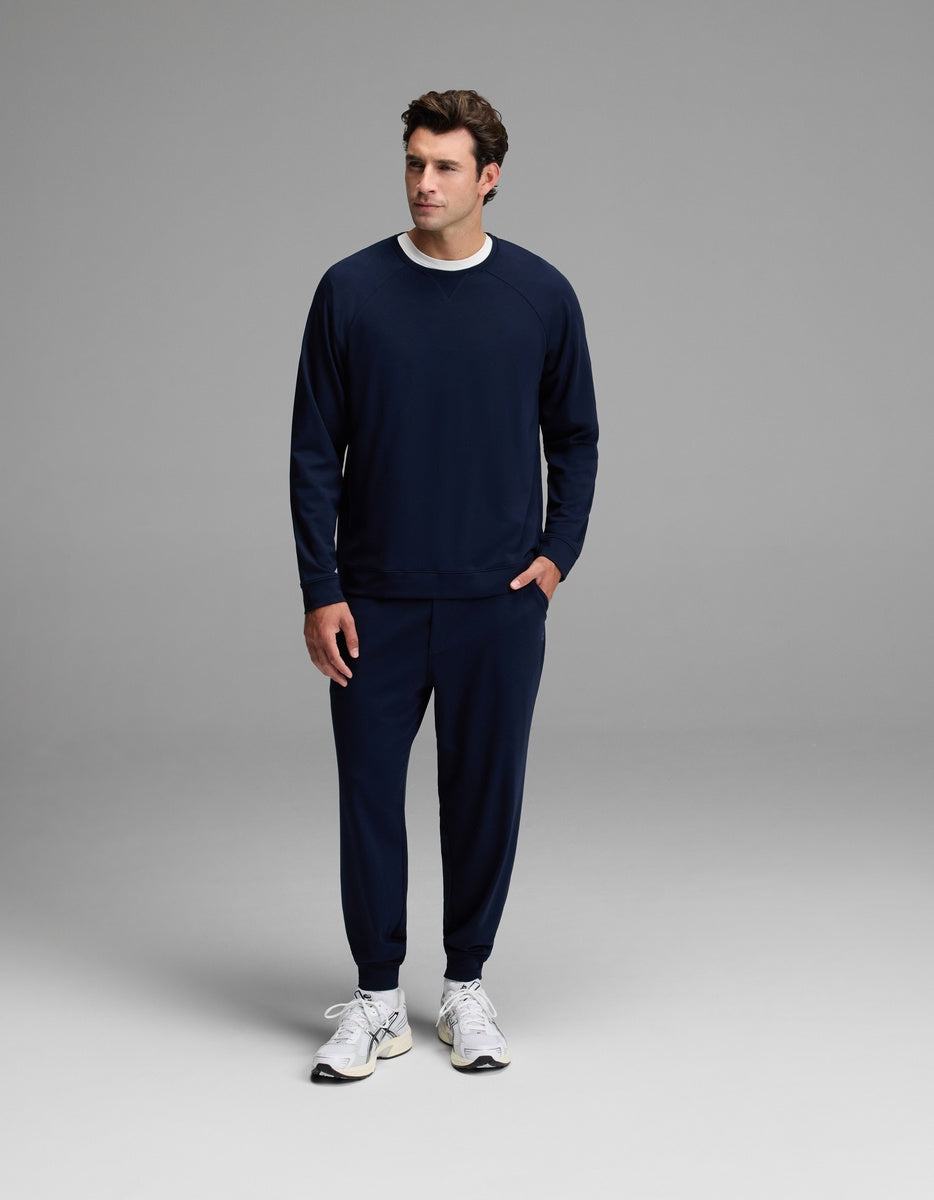 A man is seen standing against a plain gray background wearing the Cozy Earth Men’s Brushed Bamboo Jogger Set in navy blue, paired with white sneakers and a white shirt underneath. He looks slightly to his left with one hand in his pocket.