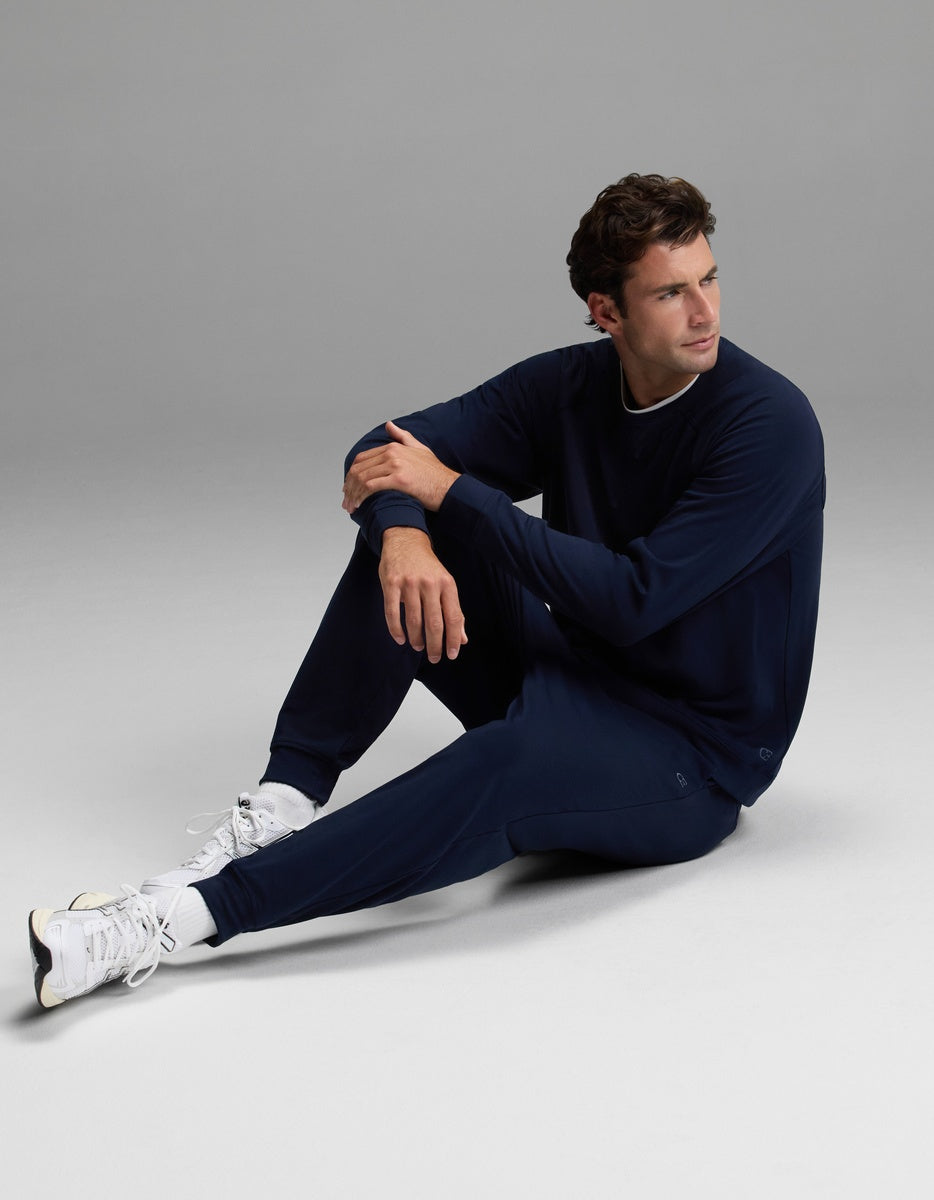 A man wearing the Cozy Earth Men’s Brushed Bamboo Jogger Set in navy blue and white sneakers sits on the floor with one leg bent and the other extended, looking off to the side against a plain gray background.