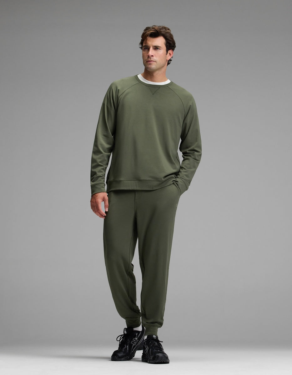 A man wearing the Cozy Earth Men’s Brushed Bamboo Jogger Set in olive green, paired with black sneakers, stands against a plain gray background, posing with one hand in his pocket and gazing slightly to the side.