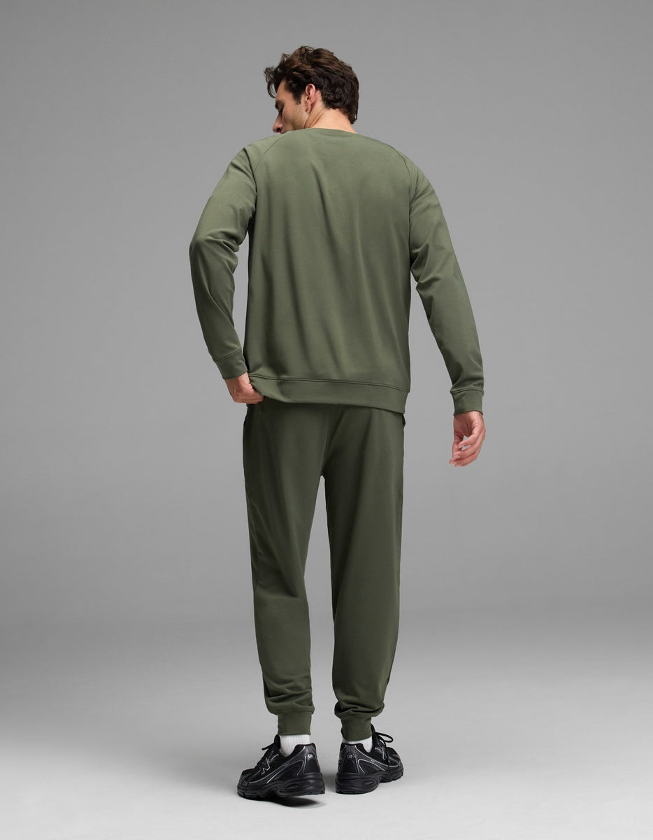 A person wearing the Cozy Earth Men’s Brushed Bamboo Jogger Set in olive green and black sneakers stands facing away, with one hand resting on their back against a plain gray background.