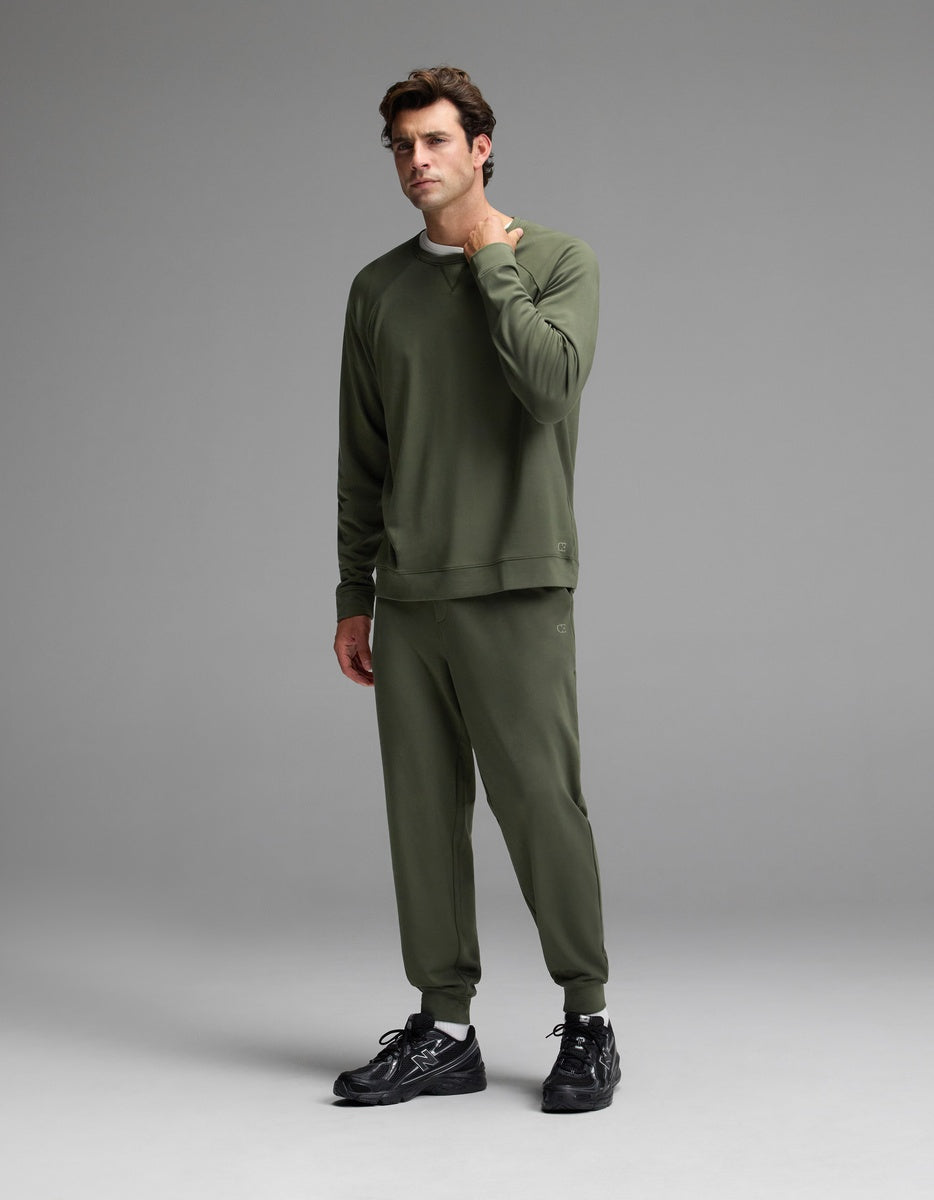 A man stands on a gray studio background, touching his neck with one hand, dressed in the Cozy Earth Men’s Brushed Bamboo Jogger Set in olive green and black sneakers.