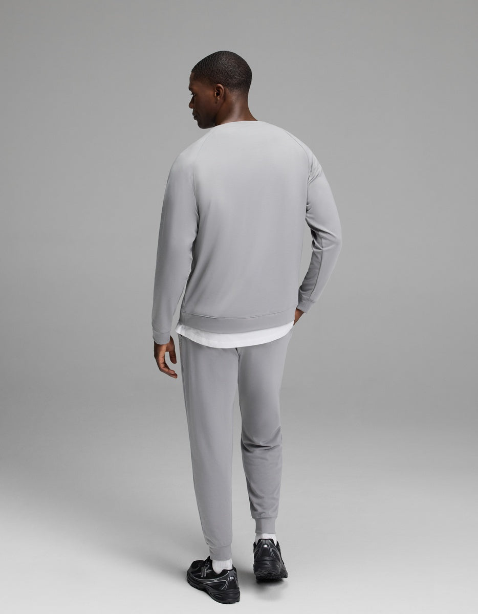 A man stands facing away, wearing the Cozy Earth Men’s Brushed Bamboo Jogger Set in light gray paired with black sneakers, his left hand resting on his hip against a plain, neutral gray background.