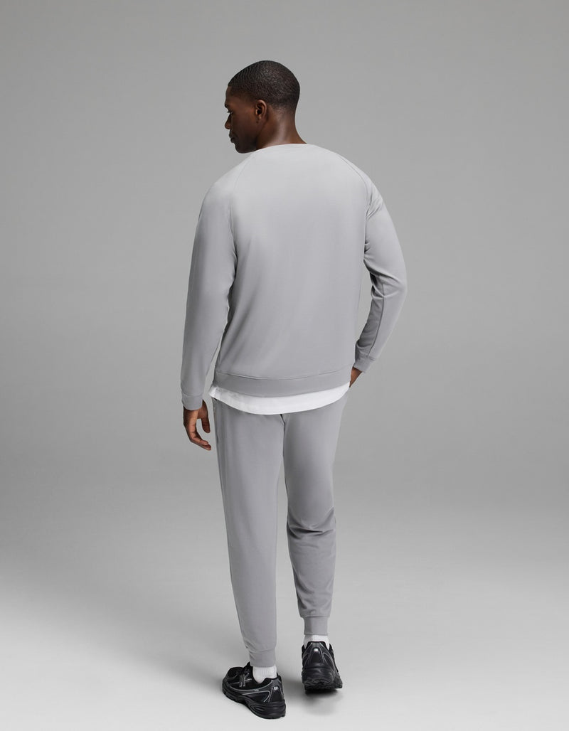A man stands facing away, wearing the Cozy Earth Men’s Brushed Bamboo Jogger Set in light gray paired with black sneakers, his left hand resting on his hip against a plain, neutral gray background.