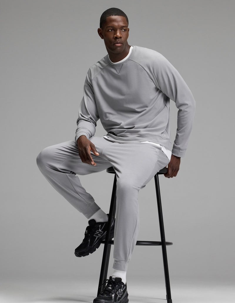A man wearing the Cozy Earth Men’s Brushed Bamboo Jogger Set in light gray sits on a tall black stool against a plain gray background. He has short hair and is wearing black athletic shoes.