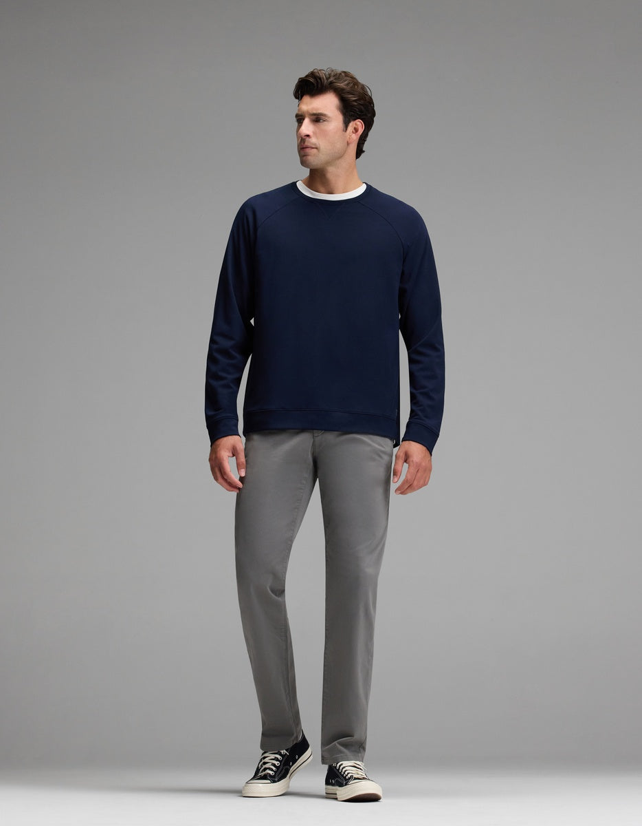 A man stands against a plain gray background, wearing the Cozy Earth Men’s Brushed Bamboo Pullover Crew in navy blue, paired with light gray pants and black sneakers with white soles, looking slightly to his left.