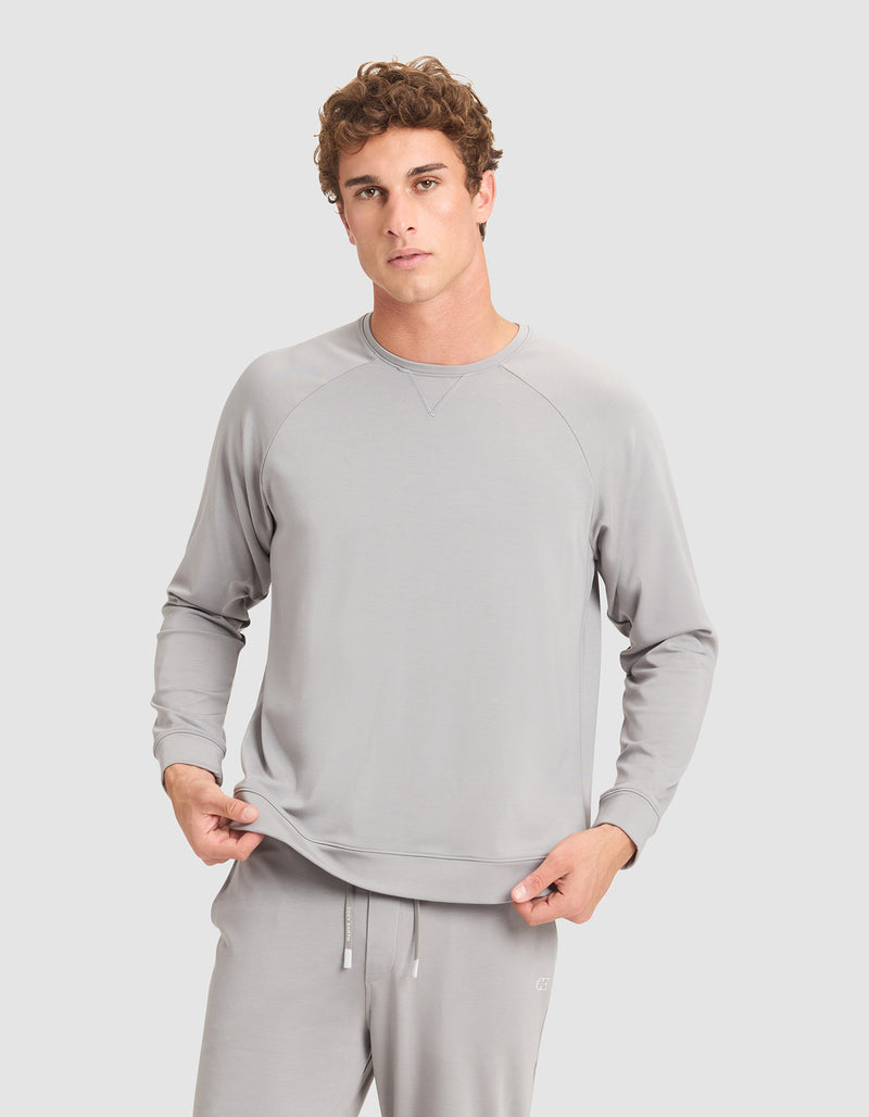 A young man with curly hair wears Cozy Earth's Men's Brushed Bamboo Pullover Crew in light gray, standing against a plain gray background and holding the hem of his sweatshirt.
