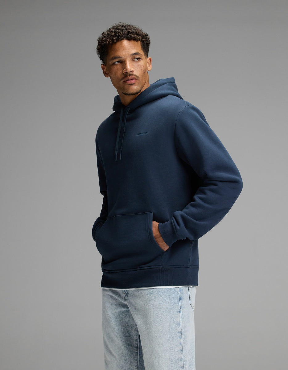 A man wearing the Cozy Earth Men’s Cityscape Hoodie in Eclipse stands against a plain gray background with his hands in the front pocket, looking to the side, and styled with light blue jeans.|Color:Eclipse