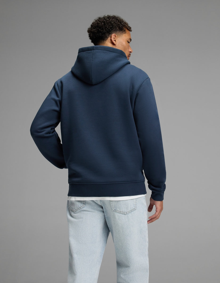 A person stands facing away, wearing the Cozy Earth Men’s Cityscape Hoodie in navy blue and light blue jeans, against a plain gray background.|Color:Eclipse