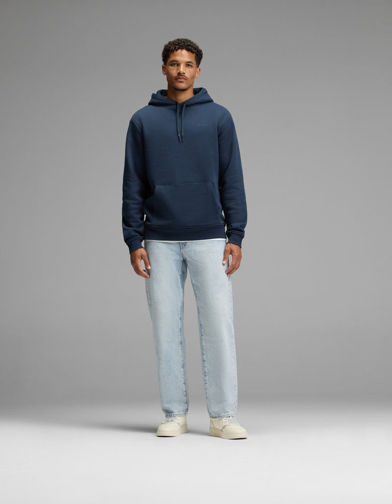 A man stands facing forward against a plain gray background, wearing the Cozy Earth Men’s Cityscape Hoodie in navy blue, paired with light blue jeans and off-white sneakers.