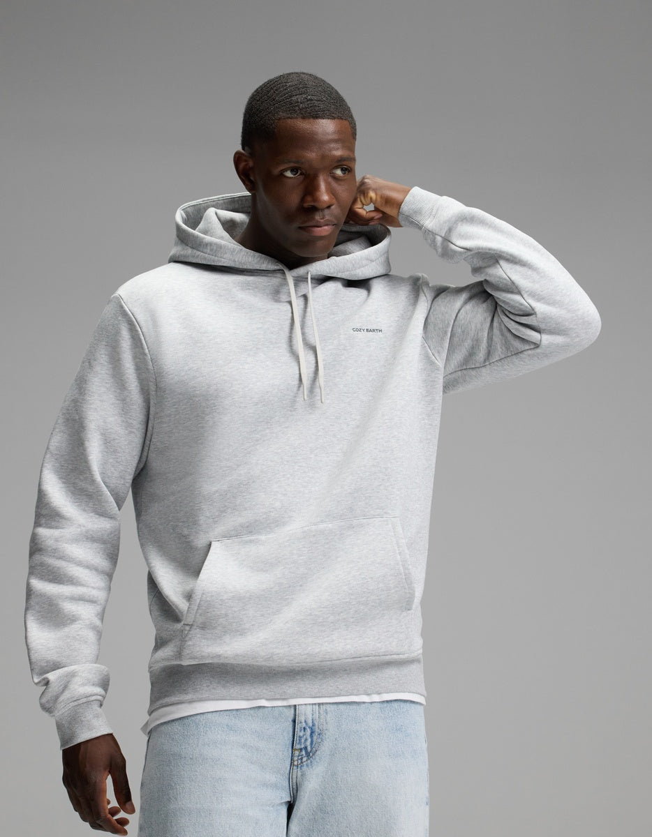 A man wearing the Cozy Earth Men’s Cityscape Hoodie in Heather Grey and light blue jeans stands against a plain gray background, with one hand touching his head and the other relaxed by his side.