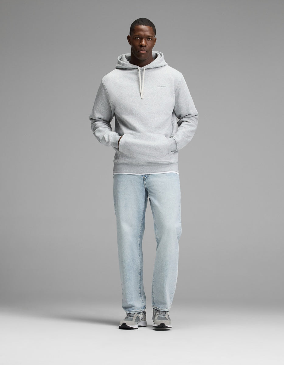 A man stands facing forward with his hands in the front pocket of a Cozy Earth Men’s Cityscape Hoodie in light gray, paired with light blue jeans and gray sneakers against a plain gray background.
