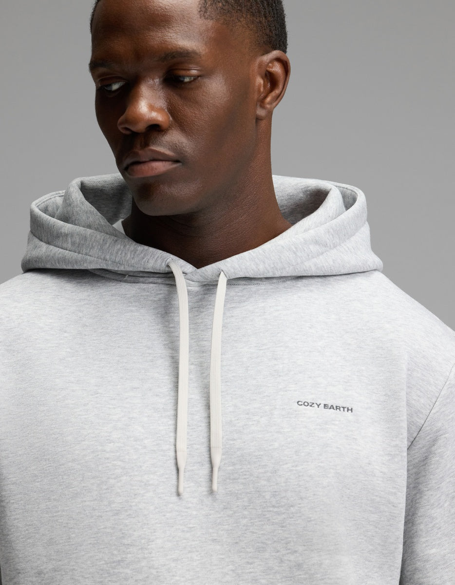 A man wearing the Cozy Earth Men’s Cityscape Hoodie in light gray, featuring white drawstrings and “COZY EARTH” text on the chest, looks down with a neutral expression against a plain gray background.