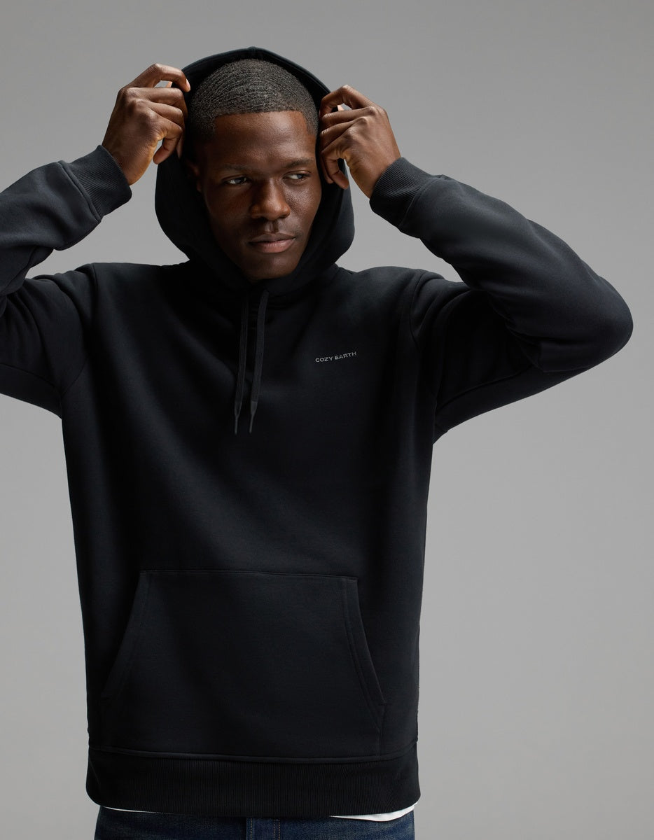 A man wears the Cozy Earth Men’s Cityscape Hoodie in Jet Black, lifting the hood over his head as he stands against a plain gray background, gazing to the side with a neutral expression.|Color:Jet Black