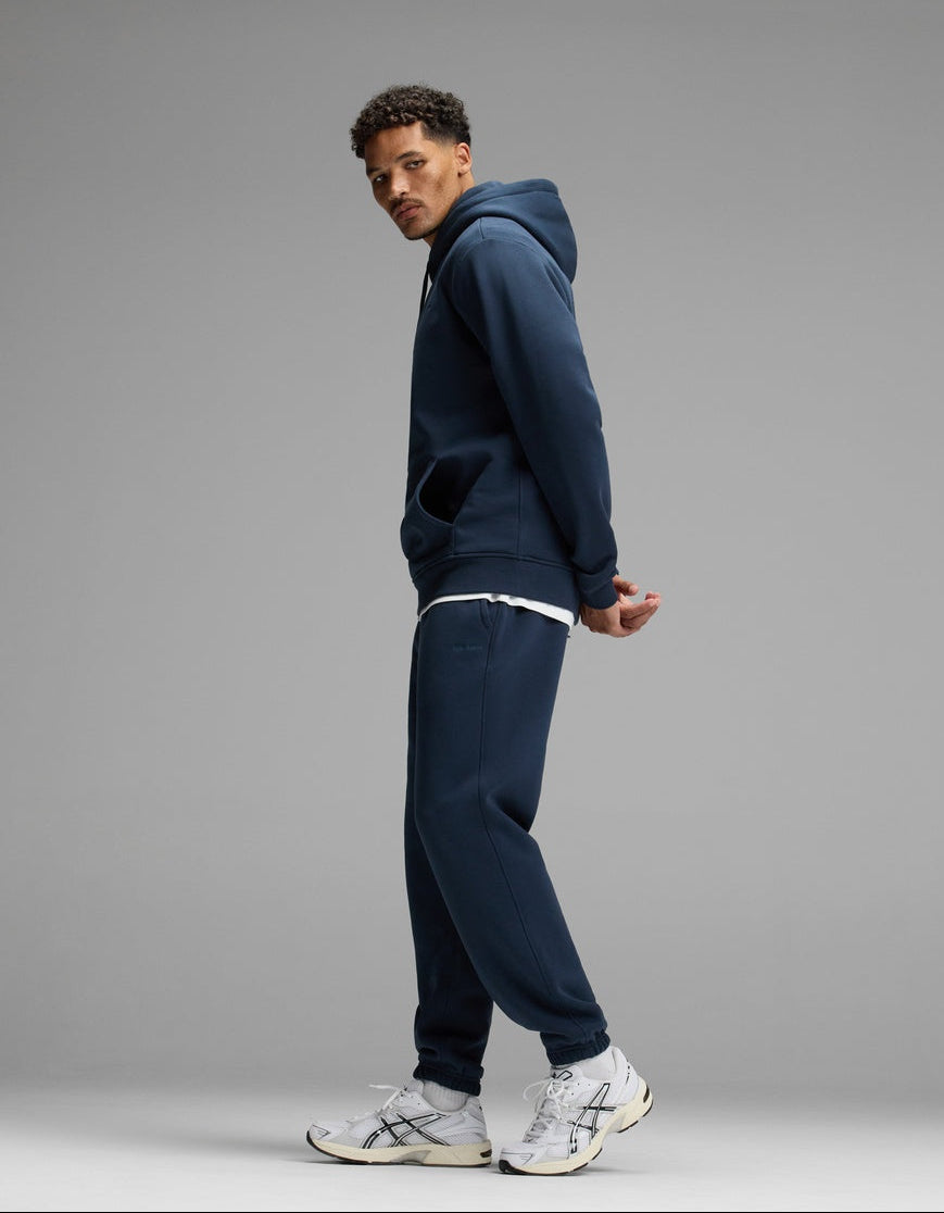 A man models the Cozy Earth Men’s Cityscape Hoodie & Sweatpant Set in navy, paired with a white T-shirt and white sneakers, standing sideways against a plain gray background with his hands behind his back and looking over his shoulder.|Color:Eclipse