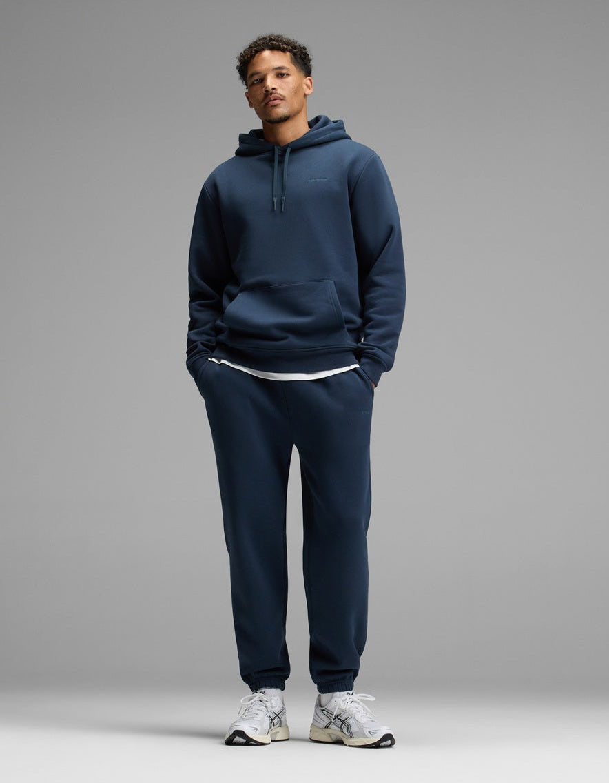 A person stands against a plain gray background, wearing the Cozy Earth Men’s Cityscape Hoodie & Sweatpant Set in Eclipse, paired with white sneakers and a white undershirt. Their hands are in their pockets and they are looking straight ahead.|Color:Eclipse