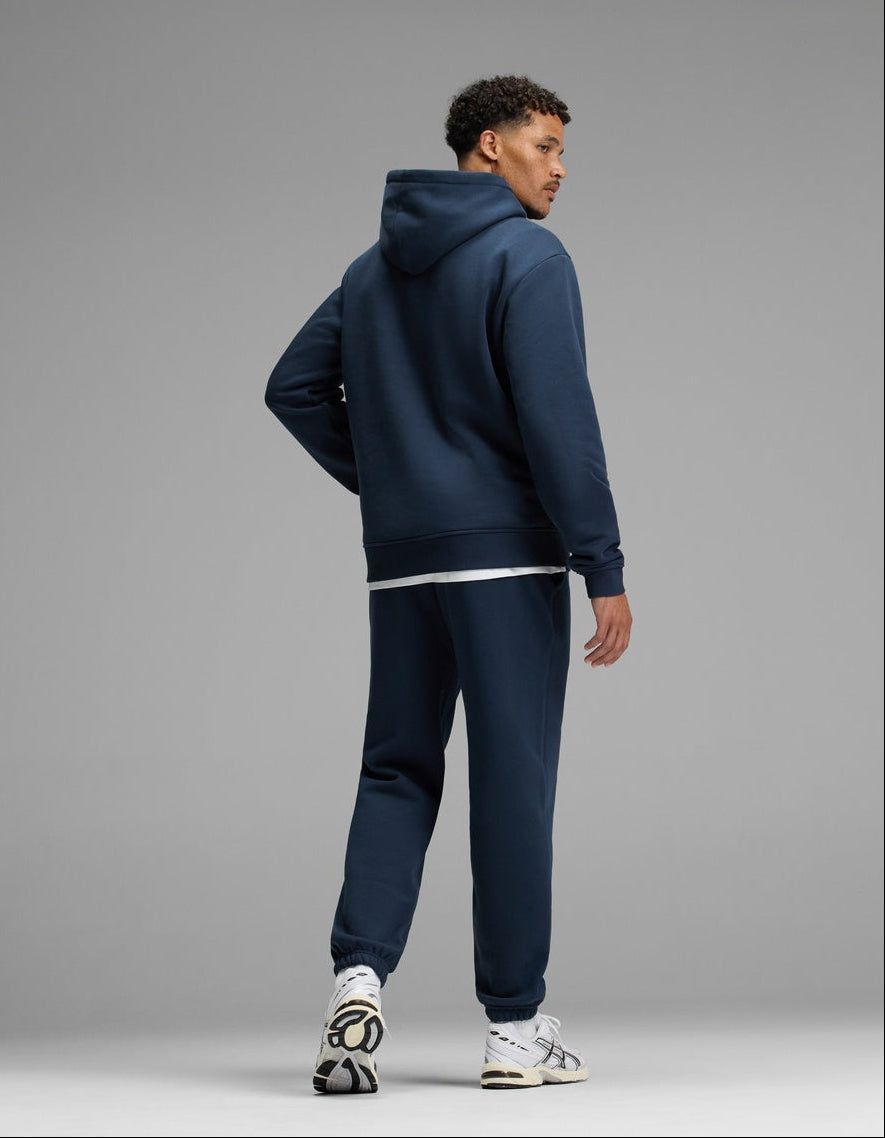 A man stands facing slightly away, wearing the Cozy Earth Men’s Cityscape Hoodie & Sweatpant Set in navy blue with white sneakers against a plain gray background.