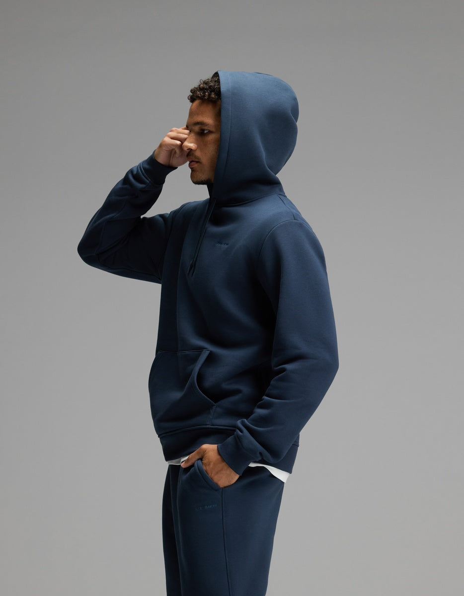 A person dressed in the Cozy Earth Men’s Cityscape Hoodie & Sweatpant Set in navy stands in profile against a plain gray background, with one hand touching their forehead.
