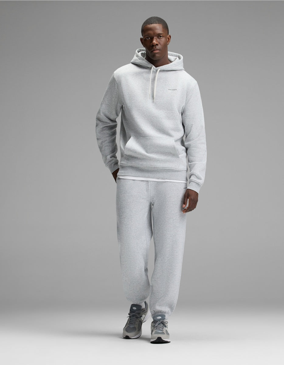 A man stands against a gray backdrop wearing the Cozy Earth Men’s Cityscape Hoodie & Sweatpant Set in Heather Grey, paired with gray athletic shoes. His left hand is in his pocket, and he faces forward with a neutral expression.