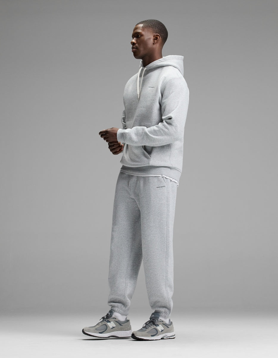 A person stands in profile against a gray background, wearing the Cozy Earth Men’s Cityscape Hoodie & Sweatpant Set paired with gray sneakers. The outfit looks casual and comfortable.