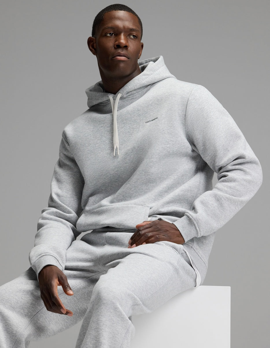 A man wearing the Cozy Earth Men’s Cityscape Hoodie & Sweatpant Set in light gray sits on a white block against a plain gray background, looking to the side with a neutral expression.