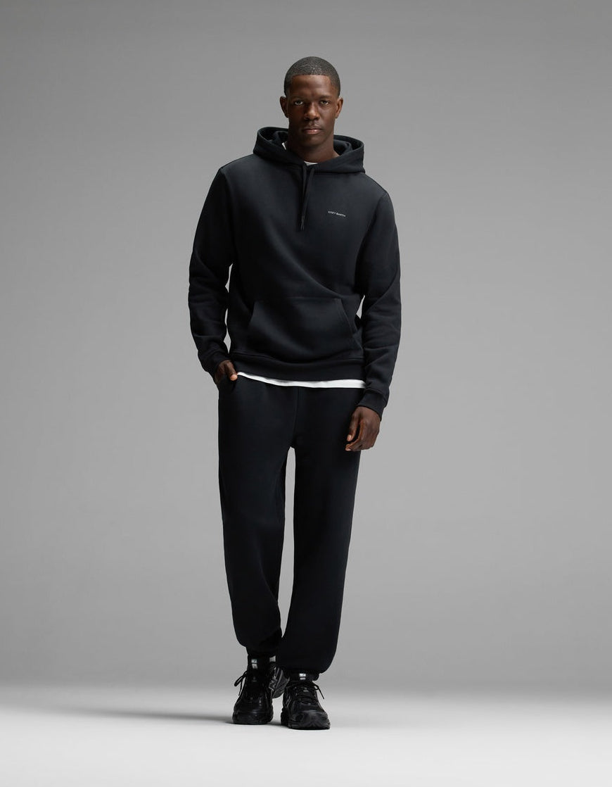 A man wearing Cozy Earth’s Men’s Cityscape Hoodie & Sweatpant Set in Jet Black stands against a plain gray background, facing the camera with a neutral expression.