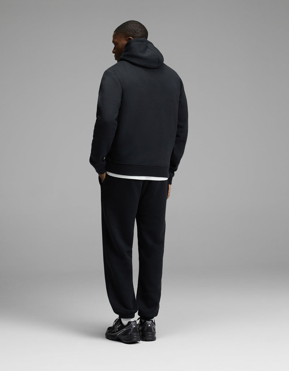 A person stands facing away, wearing the Cozy Earth Men’s Cityscape Hoodie & Sweatpant Set in black against a minimal, plain gray background.