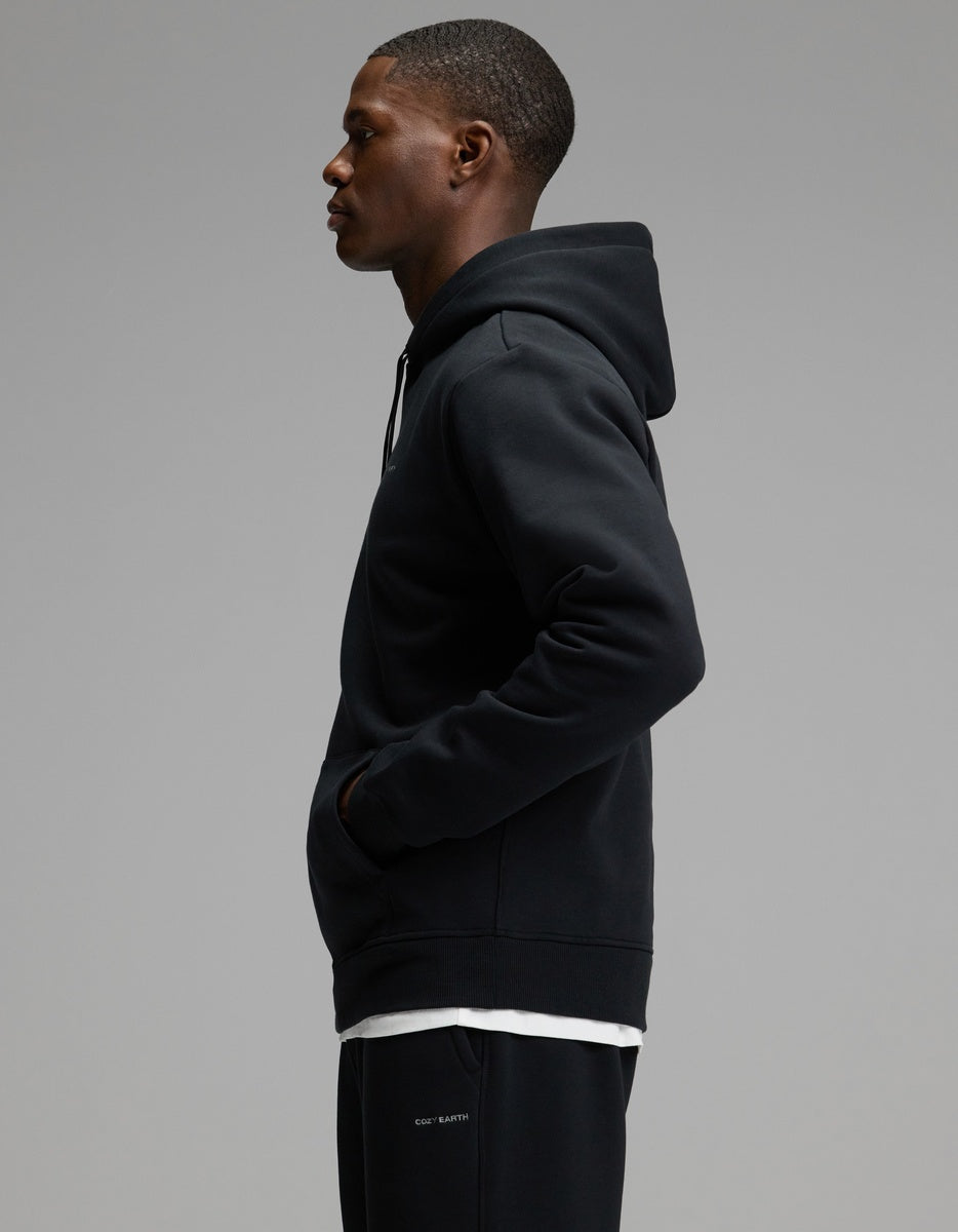 A man wearing the Cozy Earth Men’s Cityscape Hoodie & Sweatpant Set stands in profile against a plain gray background, his hands in the hoodie pocket as he looks forward.