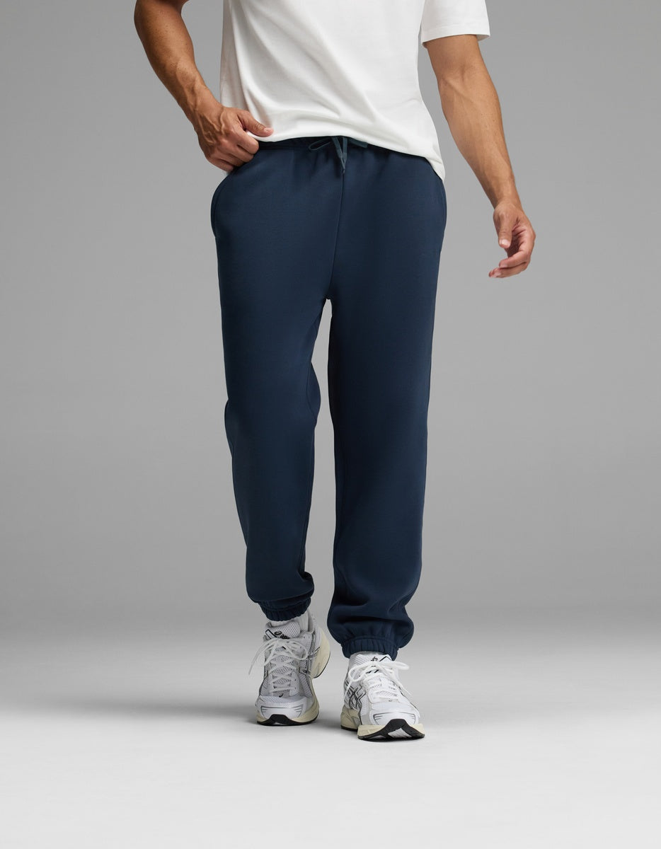 A person wearing a white T-shirt, white sneakers, and the Cozy Earth Men’s Cityscape Sweatpant in navy blue stands against a plain gray background. The image is cropped to show the individual from the shoulders down.|Color:Eclipse