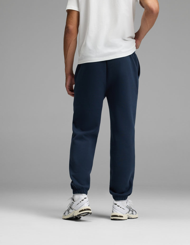 A person stands facing away, wearing a white T-shirt, Cozy Earth’s Men’s Cityscape Sweatpant in navy blue, and white sneakers. The background is plain and gray.